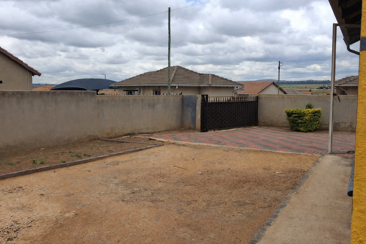 Rear yard area featuring a paved driveway section, metal gate, and boundary walls, with an unlandscaped dirt ground surface.