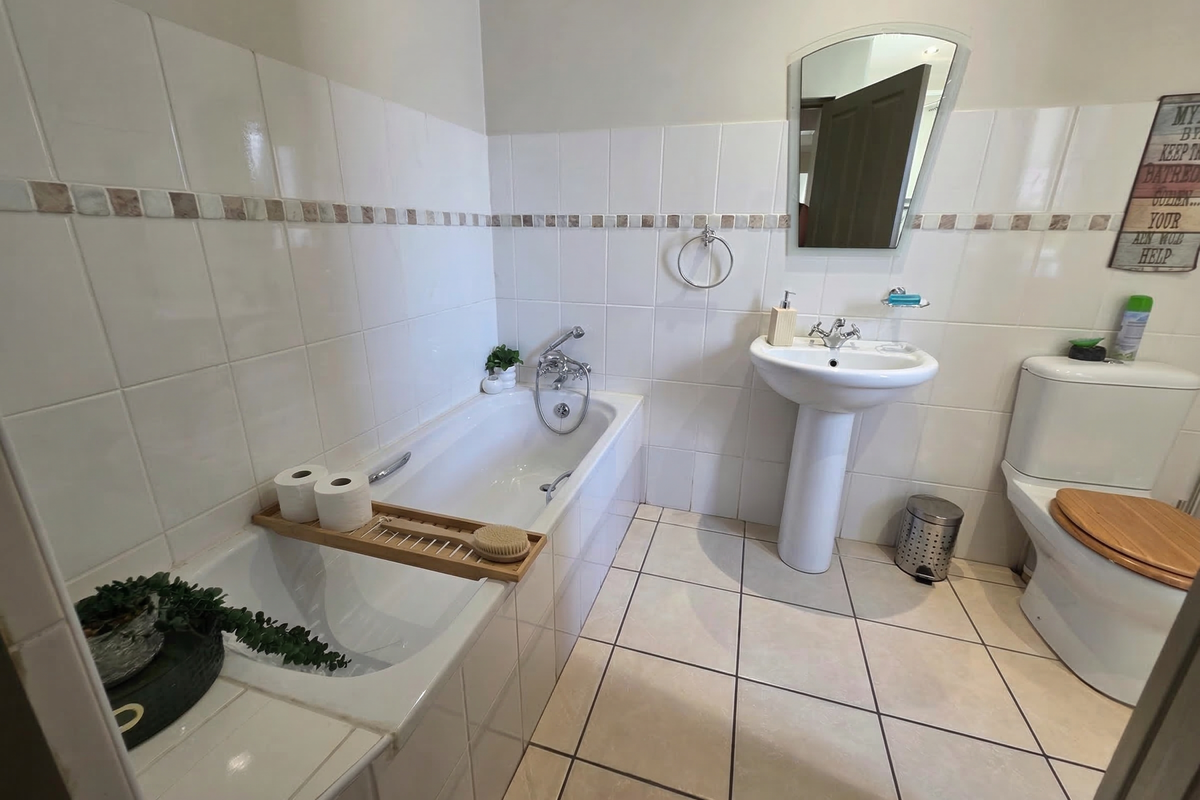 A functional bathroom featuring a built-in bathtub, pedestal basin, and toilet with neutral ceramic wall and floor tiling.