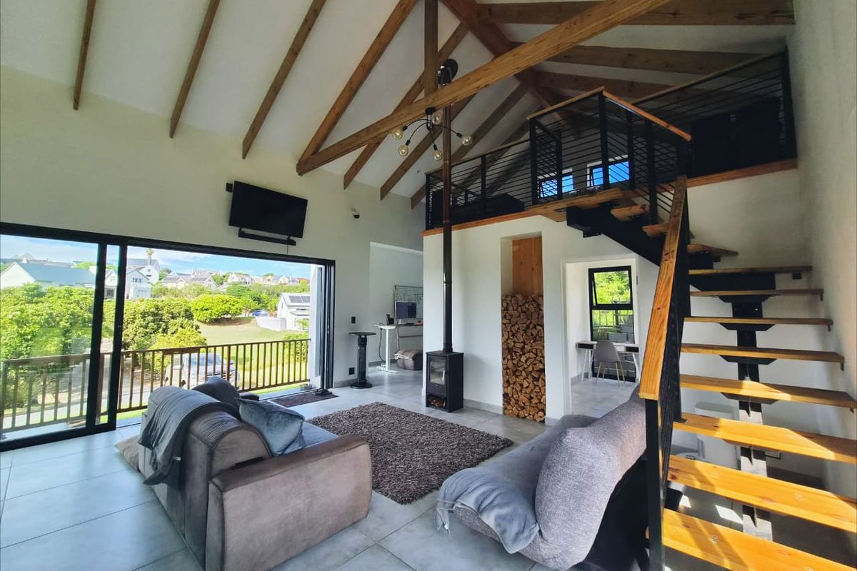 Spacious open-plan living area featuring a vaulted timber ceiling, wood-burning fireplace, and a staircase leading to a mezzanine level with large sliding doors.