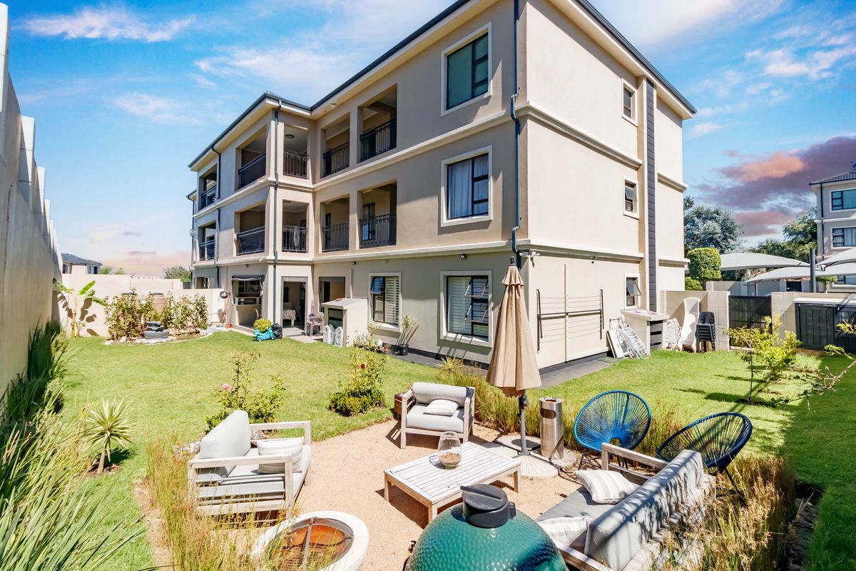 Rear view of a multi-story apartment building featuring private balconies and a well-maintained garden with a gravel patio area.