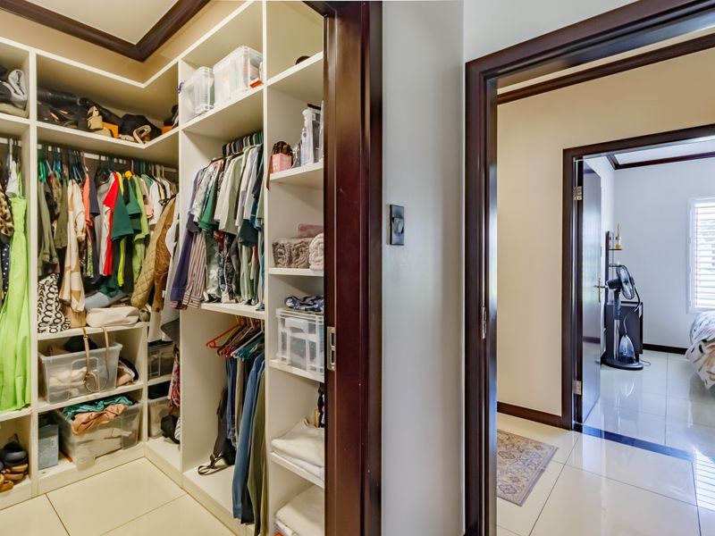 A walk-in closet with extensive built-in shelving and hanging space, leading into a bedroom with tiled floors and dark timber door frames.