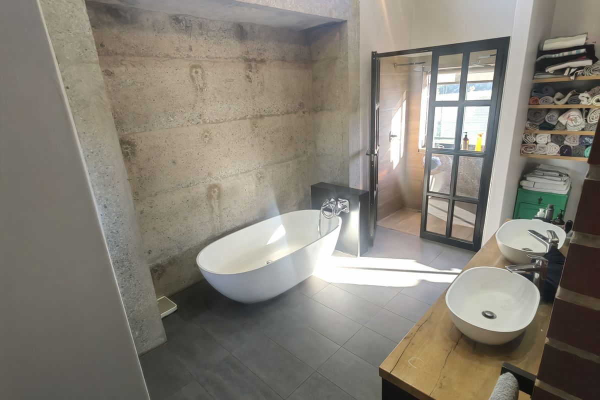 Modern bathroom featuring a freestanding bathtub, exposed concrete feature wall, double vessel sinks on a timber vanity, and a black-framed glass shower enclosure.