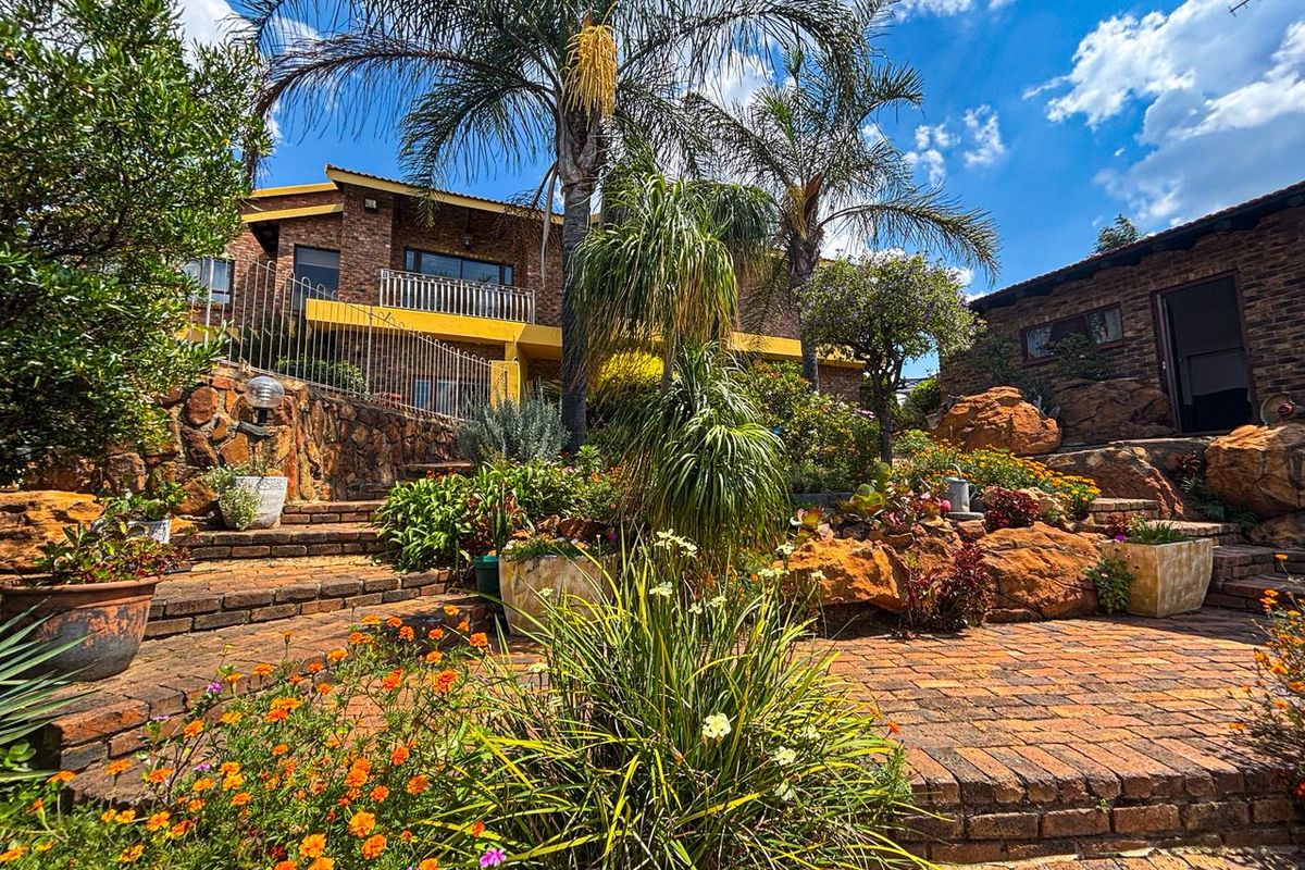 A multi-level brick residence featuring tiered landscaping, stone retaining walls, paved pathways, and a balcony overlooking a lush, established garden.