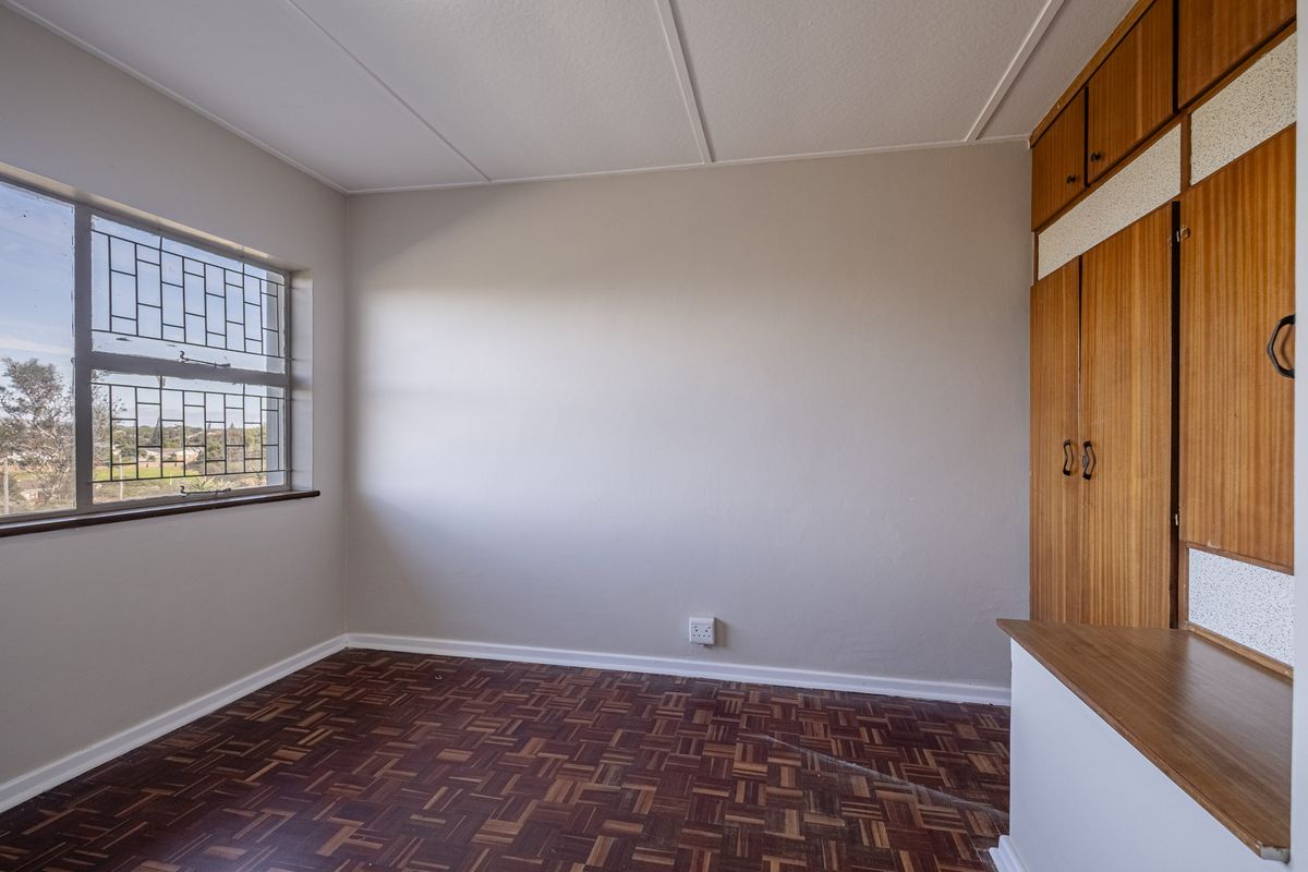 A bright bedroom featuring classic parquet flooring, built-in wooden cabinetry with integrated desk space, and a window fitted with security bars.