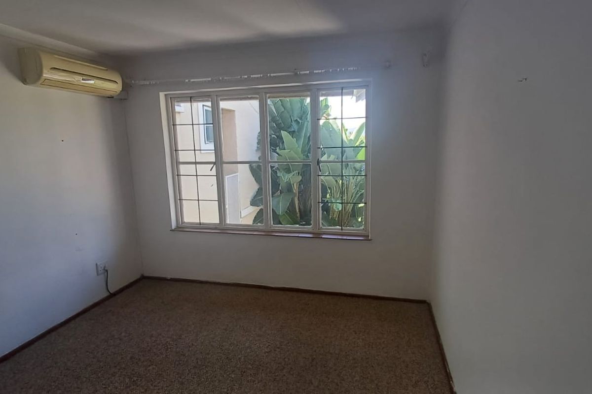 An empty bedroom featuring carpet flooring, a wall-mounted air conditioning unit, and a large window with a garden view.