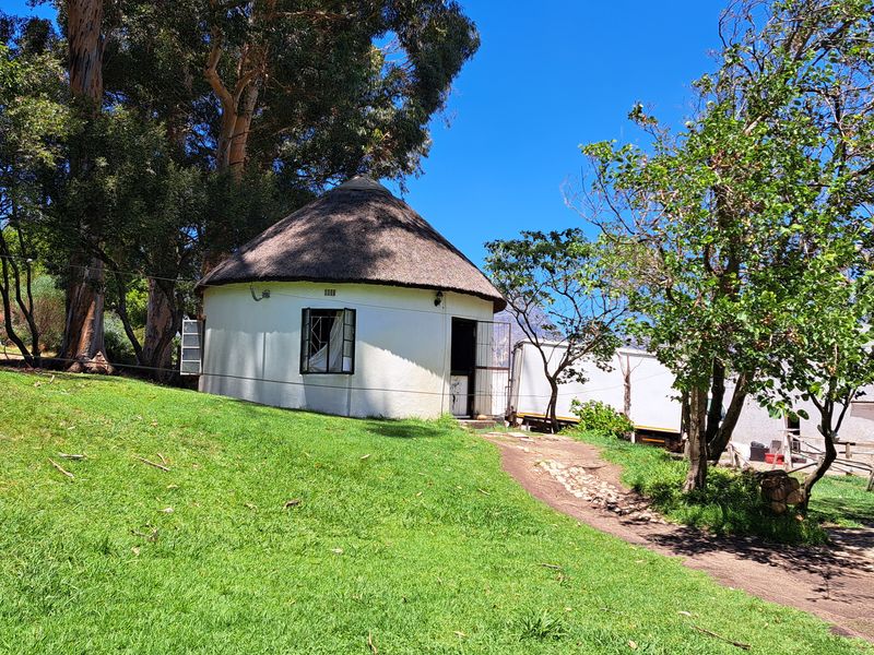 A circular cottage with a traditional thatch roof situated on a sloped, grassy garden area on the farm property.