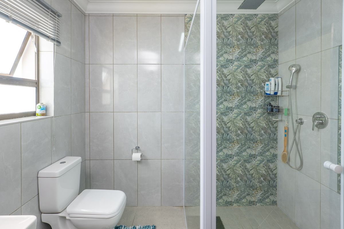 A modern bathroom featuring a glass-enclosed shower with decorative leaf-patterned tiling, a standard toilet, and neutral grey wall tiles.