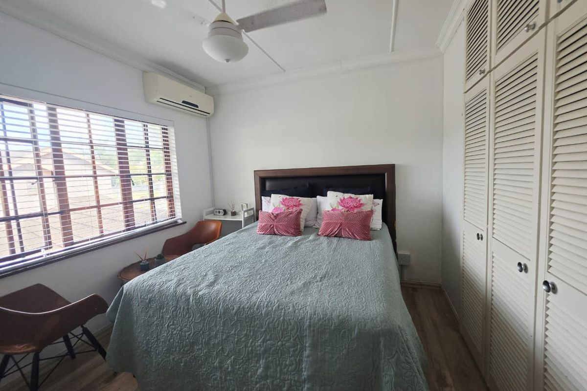 A well-lit bedroom featuring built-in louvered cupboards, laminate flooring, a ceiling fan, and a wall-mounted air conditioning unit.