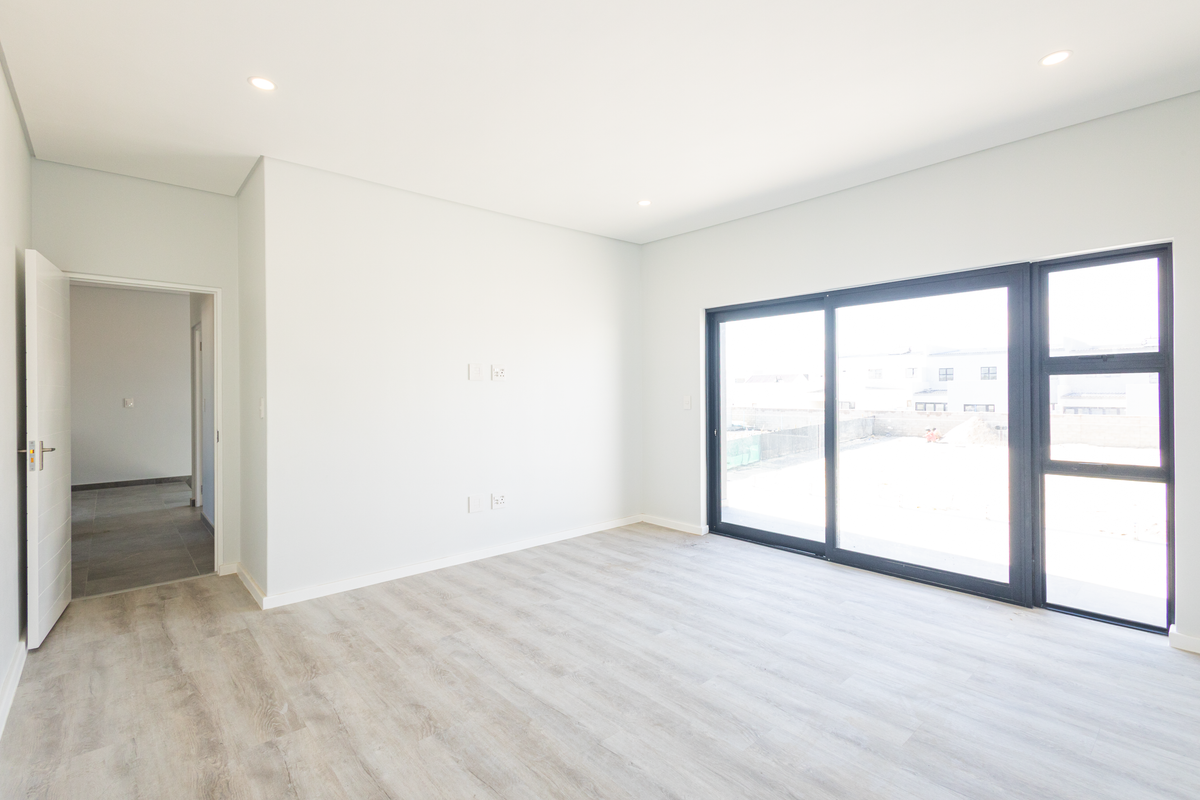 A bright, modern, and empty bedroom featuring light-toned laminate flooring, recessed ceiling lighting, and a large sliding glass door providing natural light.