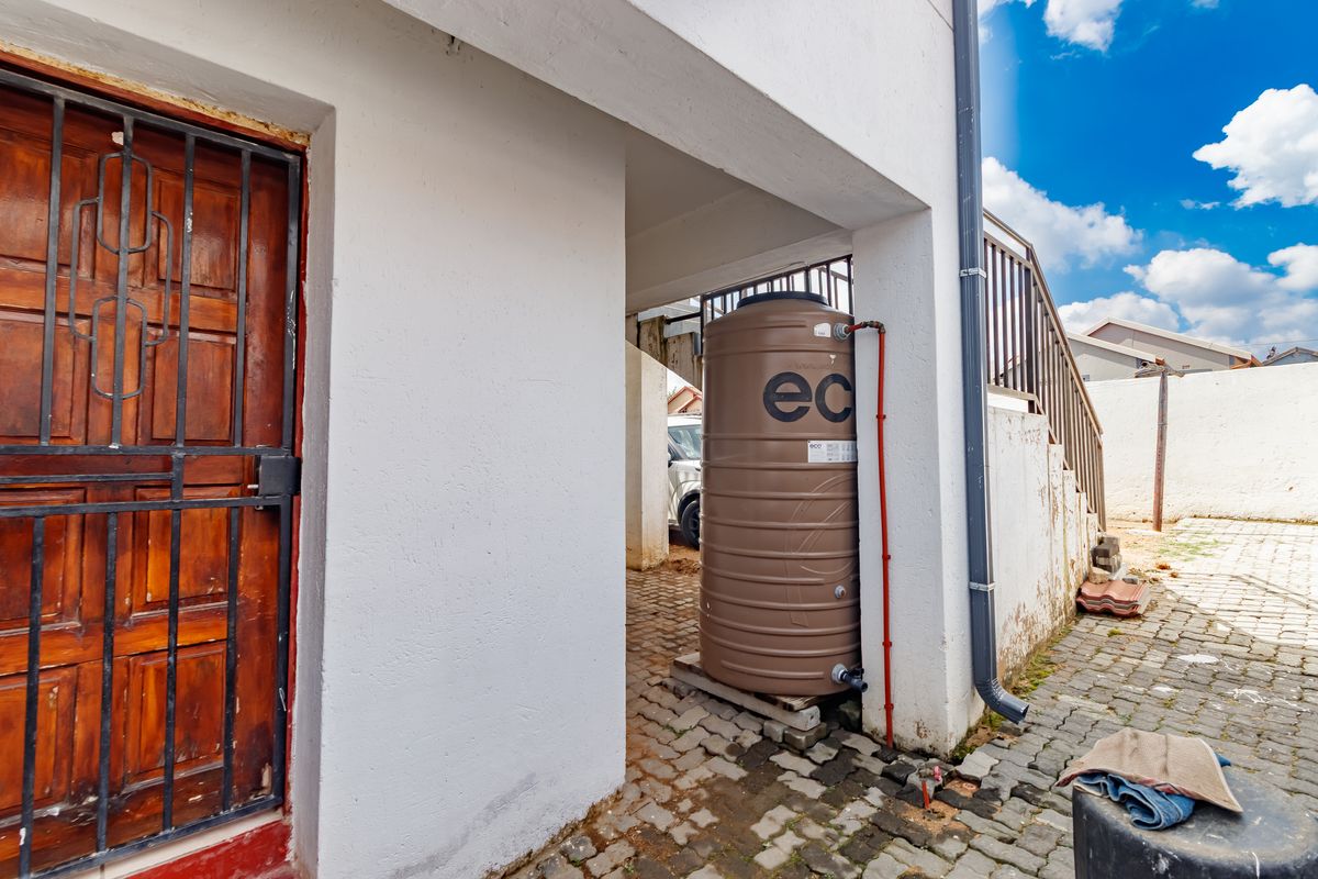Rear exterior area featuring a water storage tank, paved ground, and access to a staircase with a security gate.