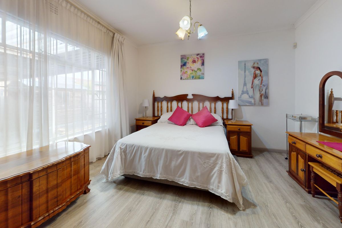 A well-lit bedroom featuring light-toned laminate flooring, a large window with curtain tracks, and a central ceiling light fixture.