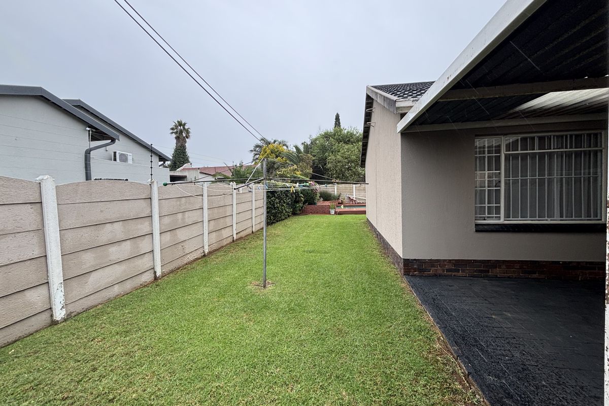 A rear garden view featuring a lawn, a covered patio area, and a precast concrete boundary wall with security bars on the house windows.