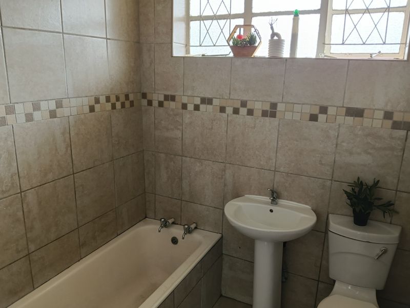 A functional bathroom featuring a pedestal basin, built-in bathtub, and toilet, finished with neutral wall tiling and a window with security bars.