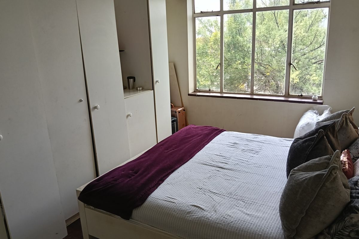 A bedroom featuring built-in cupboards, parquet flooring, and a large window providing natural light and a leafy outlook.