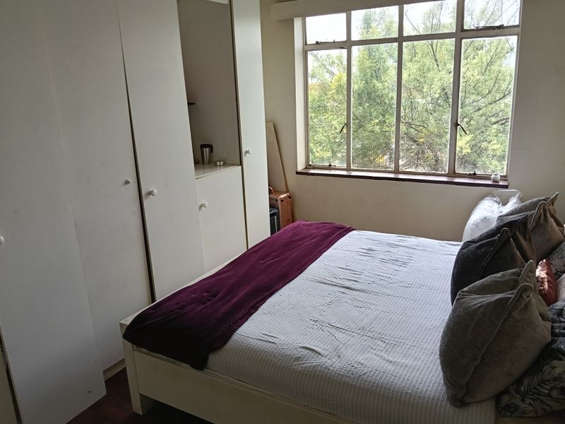A bedroom featuring built-in cupboards, parquet flooring, and a large window providing natural light and a leafy outlook.