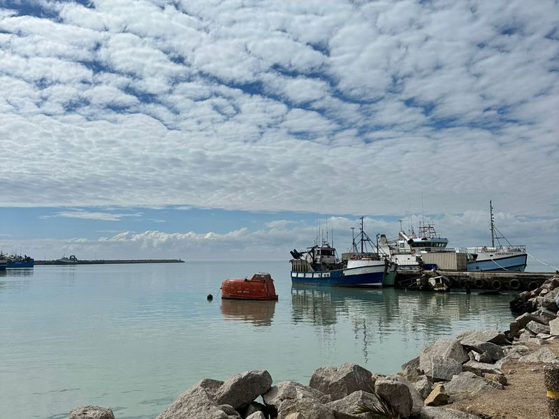 A scenic view of a harbour with fishing vessels moored at a jetty, framed by a rocky shoreline under a cloudy sky.
