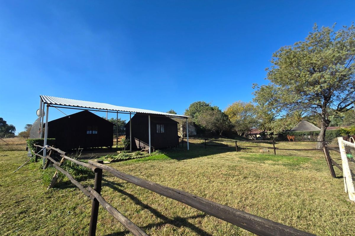 Spacious outdoor area featuring timber outbuildings sheltered by a corrugated metal carport, enclosed by rustic post and rail fencing on a small holding.