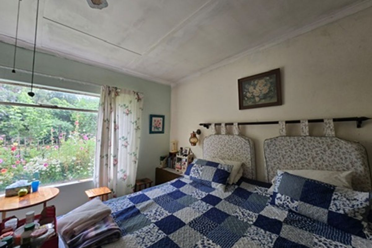 A bedroom featuring a large window with garden views and a ceiling fan. The room shows signs of wear, including visible ceiling panel cracks.