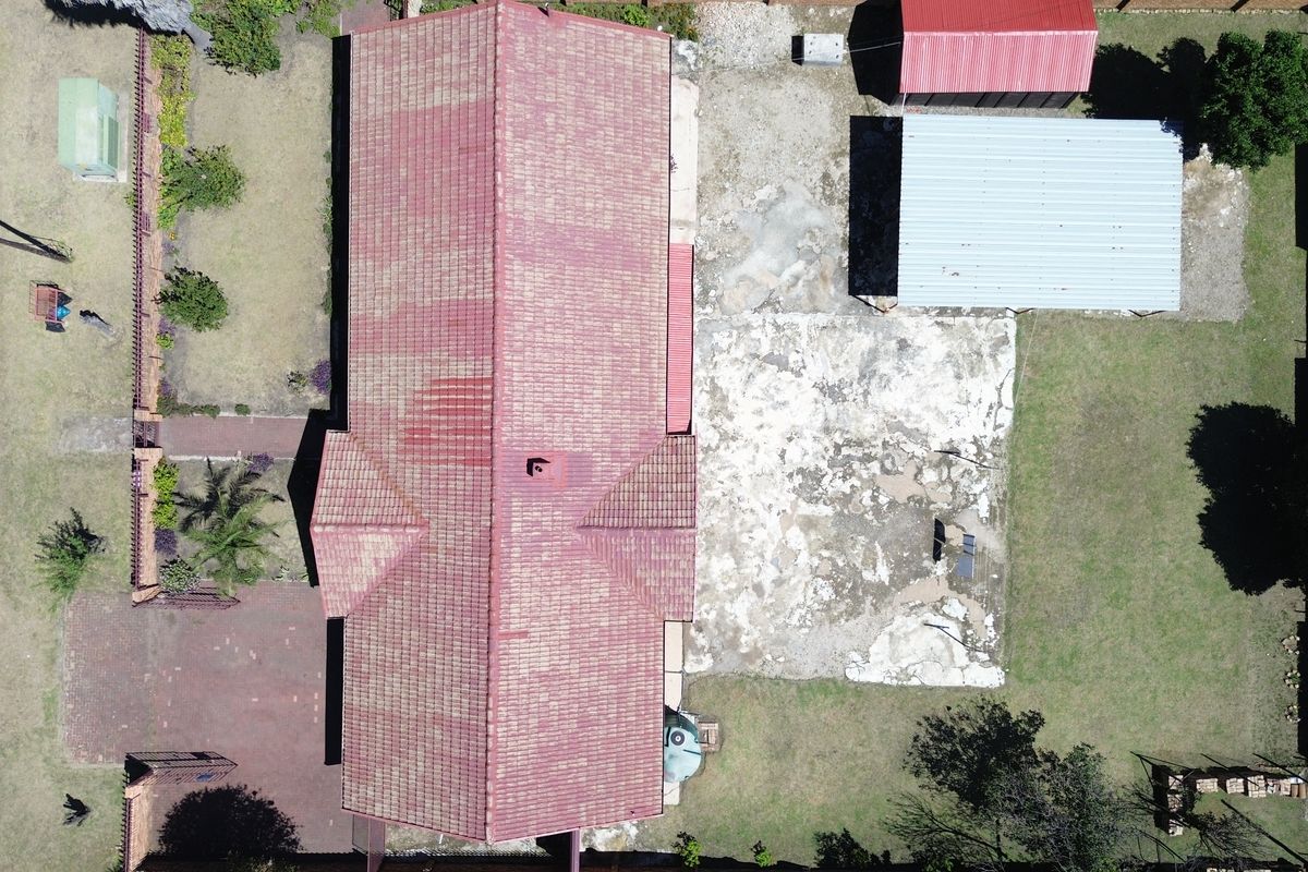 Aerial view of the property showing the main house with a tiled roof, secondary metal structures, paved driveway, and a large concrete yard area.