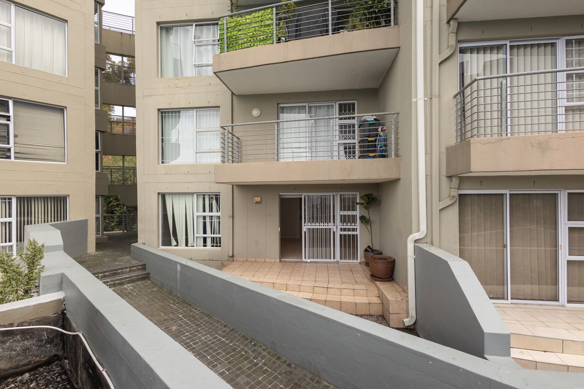 Exterior view of an apartment building featuring ground-floor tiled patio access, upper-level balconies with metal railings, and paved walkways.