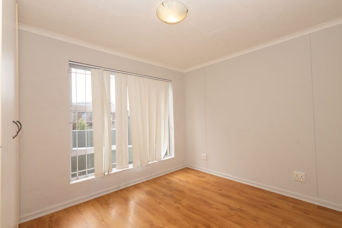 An empty bedroom featuring laminate flooring, a built-in cupboard, and a window fitted with security bars and vertical blinds.