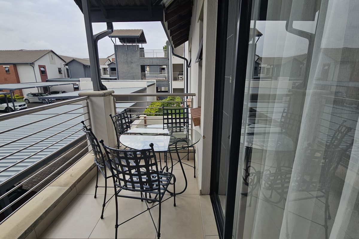 A covered balcony featuring tiled flooring and a stainless steel balustrade, offering an elevated view of the apartment complex.