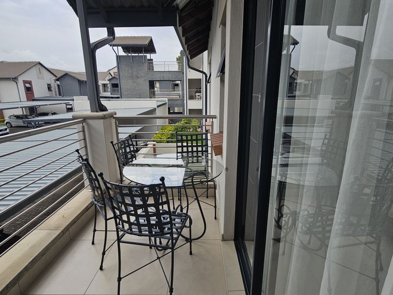 A covered balcony featuring tiled flooring and a stainless steel balustrade, offering an elevated view of the apartment complex.