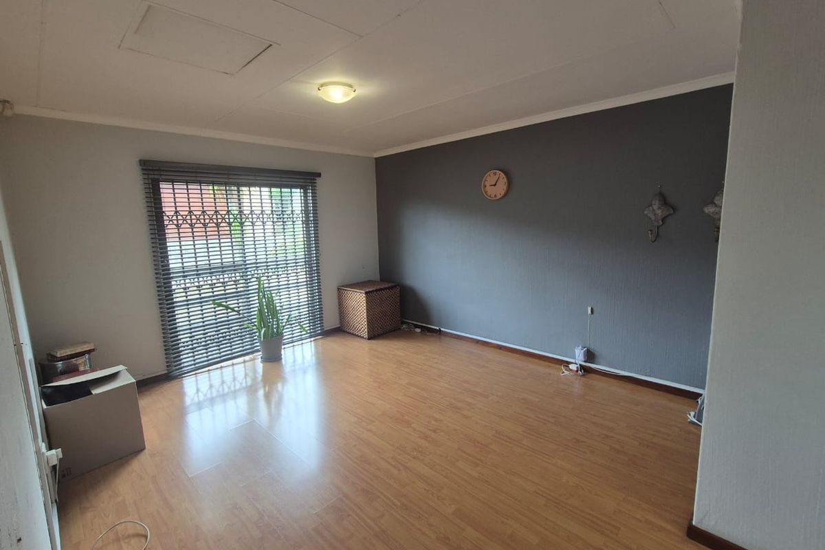 A spacious living room featuring laminate wood flooring, a feature wall, and a large window fitted with a security gate and blinds.