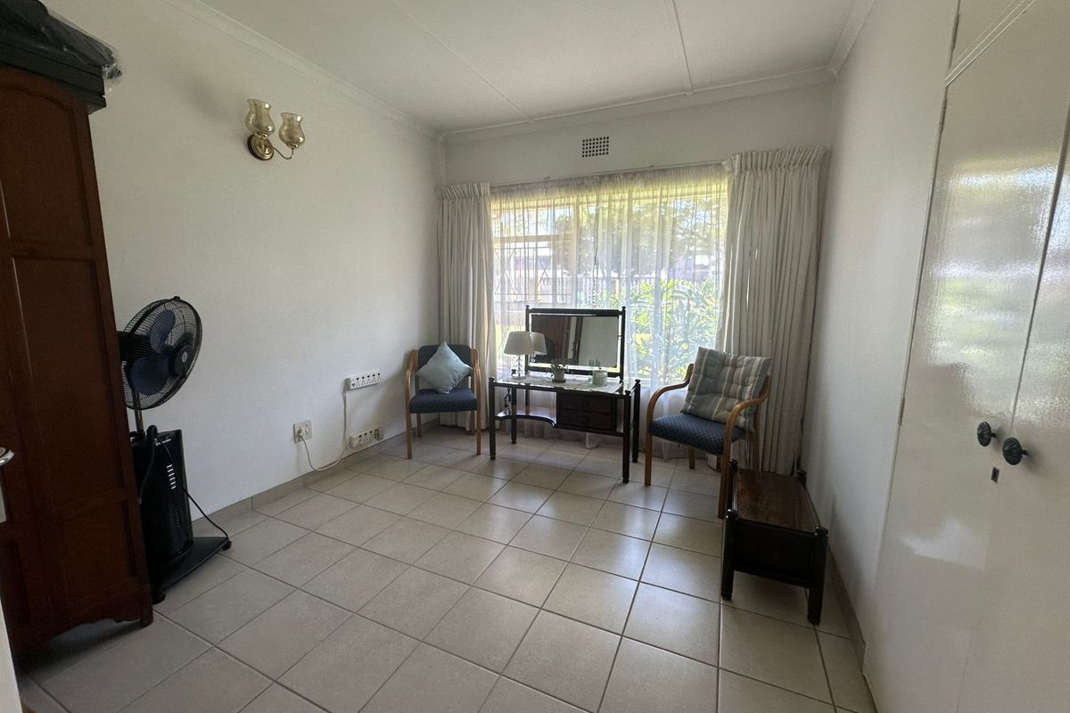 A functional bedroom featuring tiled flooring, a built-in cupboard, and a large window with security bars, offering a bright and neutral living space.
