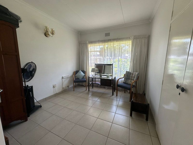A functional bedroom featuring tiled flooring, a built-in cupboard, and a large window with security bars, offering a bright and neutral living space.