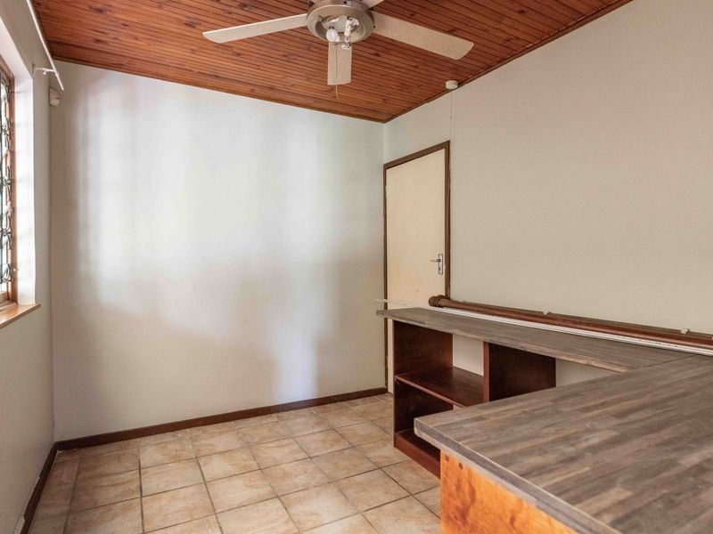 A functional office space featuring a timber ceiling, ceiling fan, tiled flooring, and a built-in desk with security bars on the window.