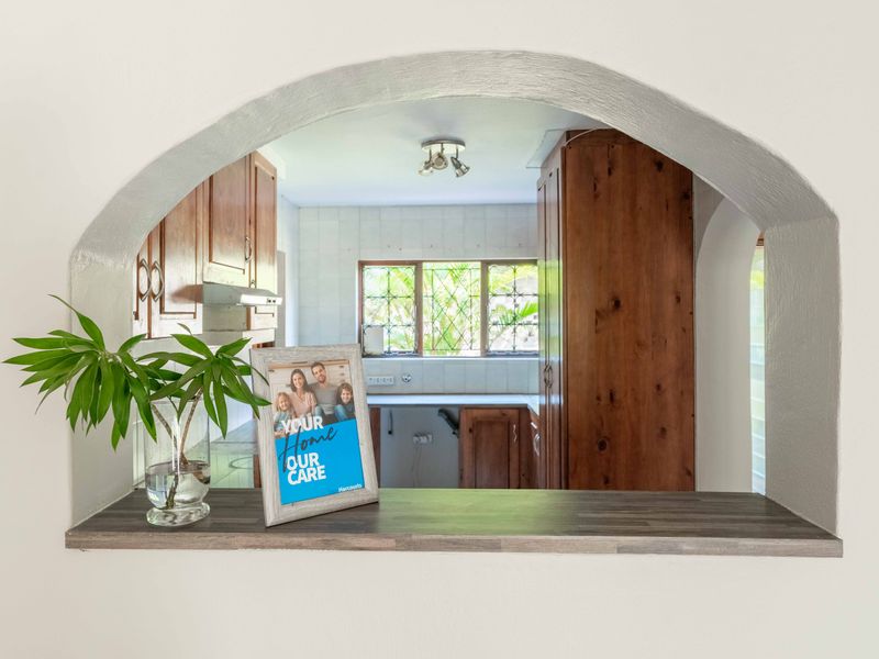 A view through a character archway into a kitchen featuring wooden cabinetry, tiled backsplash, and a window with security bars.