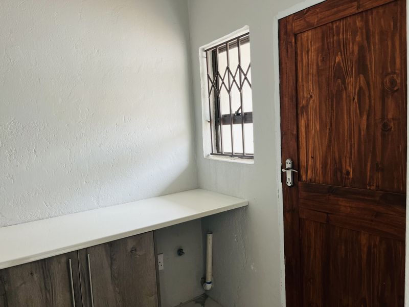 A functional laundry area featuring built-in cabinetry, a countertop, and plumbing connections, finished with a wooden door and a window with security bars.