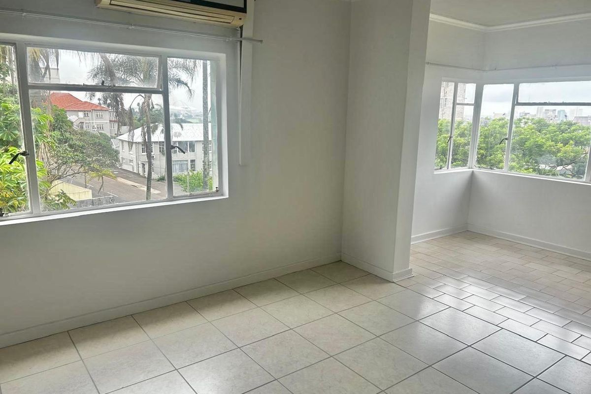 A bright, empty living area featuring large windows, tiled flooring, and a wall-mounted air conditioning unit, offering a versatile space with natural light.