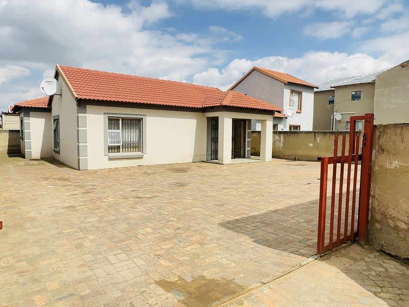 Front view of a single-story house featuring a tiled roof, paved driveway, and secure gated entrance with perimeter walling.
