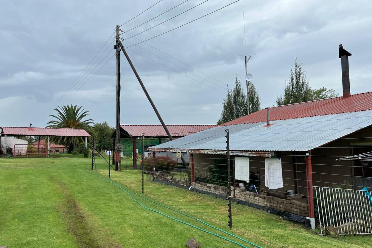 Exterior view of farm outbuildings featuring corrugated metal roofing, electric fencing, and expansive lawn areas under an overcast sky.