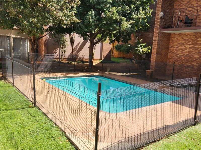A communal swimming pool area featuring a paved surround, safety fencing, and mature trees, set against a brick apartment building.