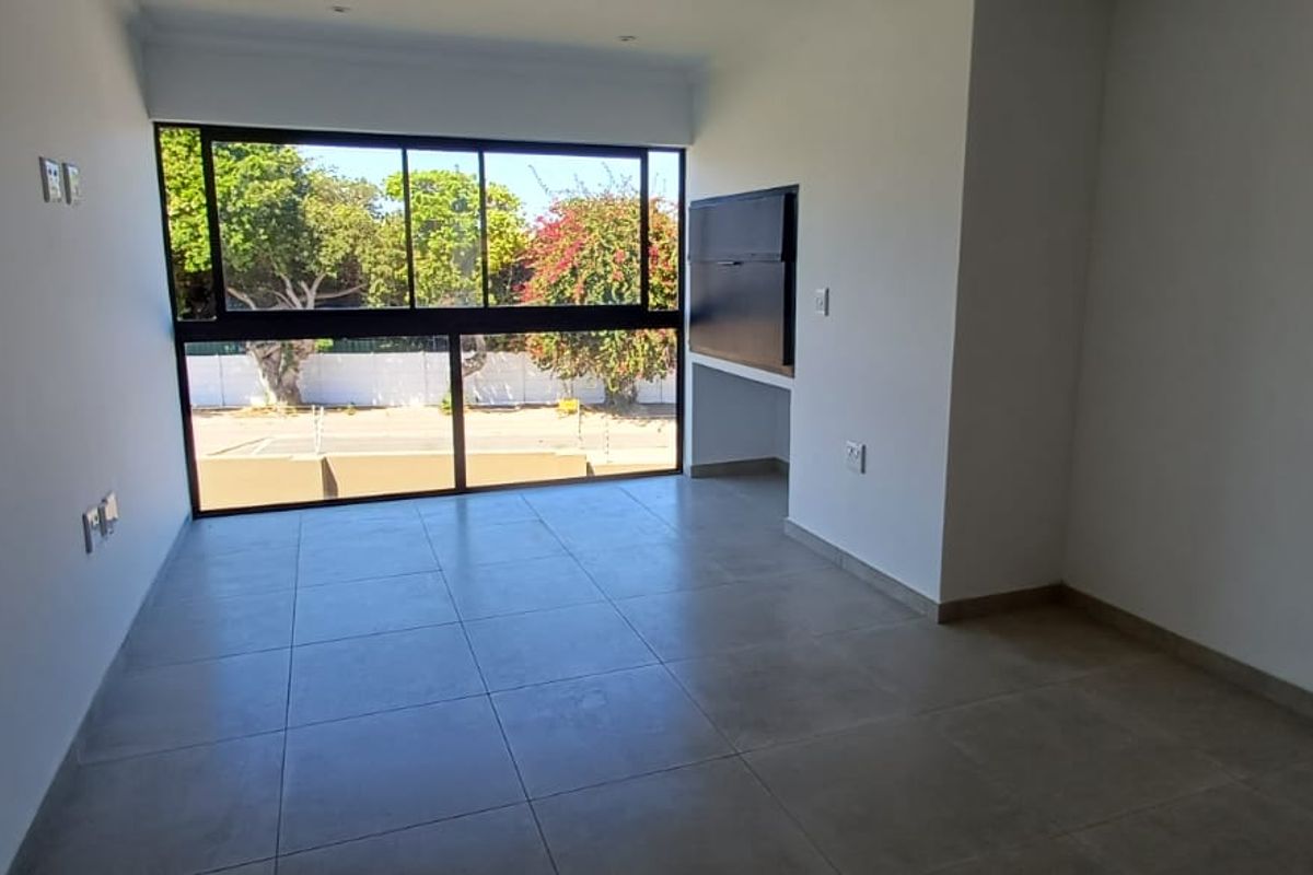 A bright, empty living area featuring large format floor tiles and expansive black-framed windows providing natural light and outdoor views.