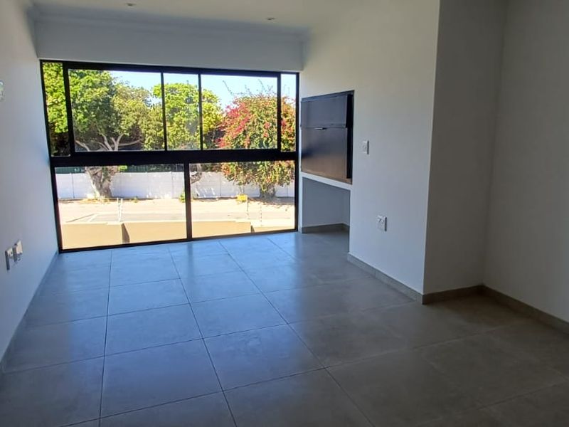 A bright, empty living area featuring large format floor tiles and expansive black-framed windows providing natural light and outdoor views.