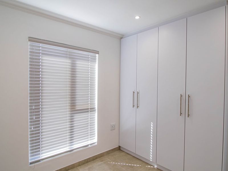 A clean, modern bedroom featuring white built-in cupboards, tiled flooring, and venetian blinds, offering a bright and neutral living space.