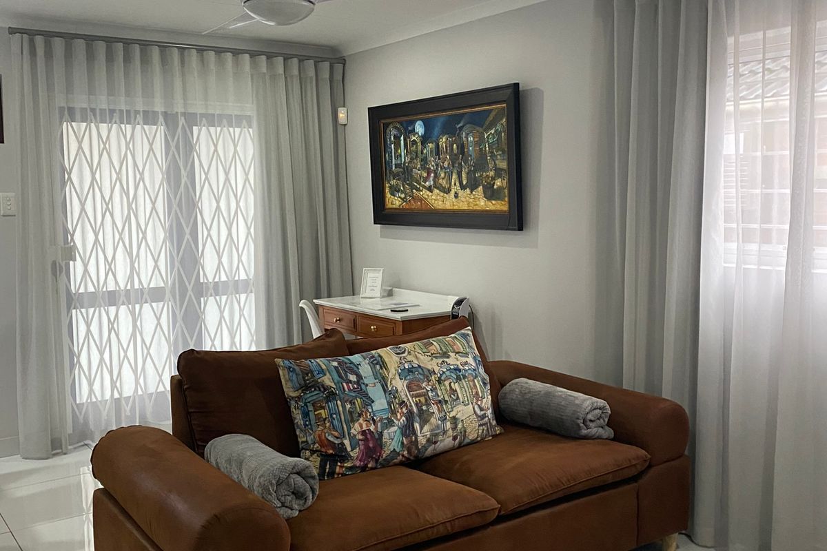A well-maintained living room featuring neutral tiled flooring, a modern ceiling fan, and large windows with fitted curtain tracks and security gates.