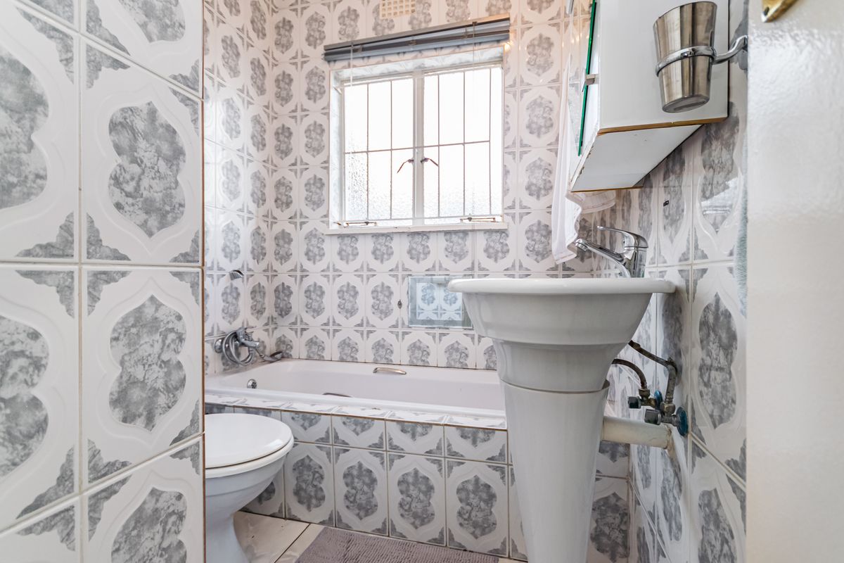 A compact bathroom featuring a pedestal basin, bathtub, and toilet, finished with dated patterned wall tiles and a window with security bars.