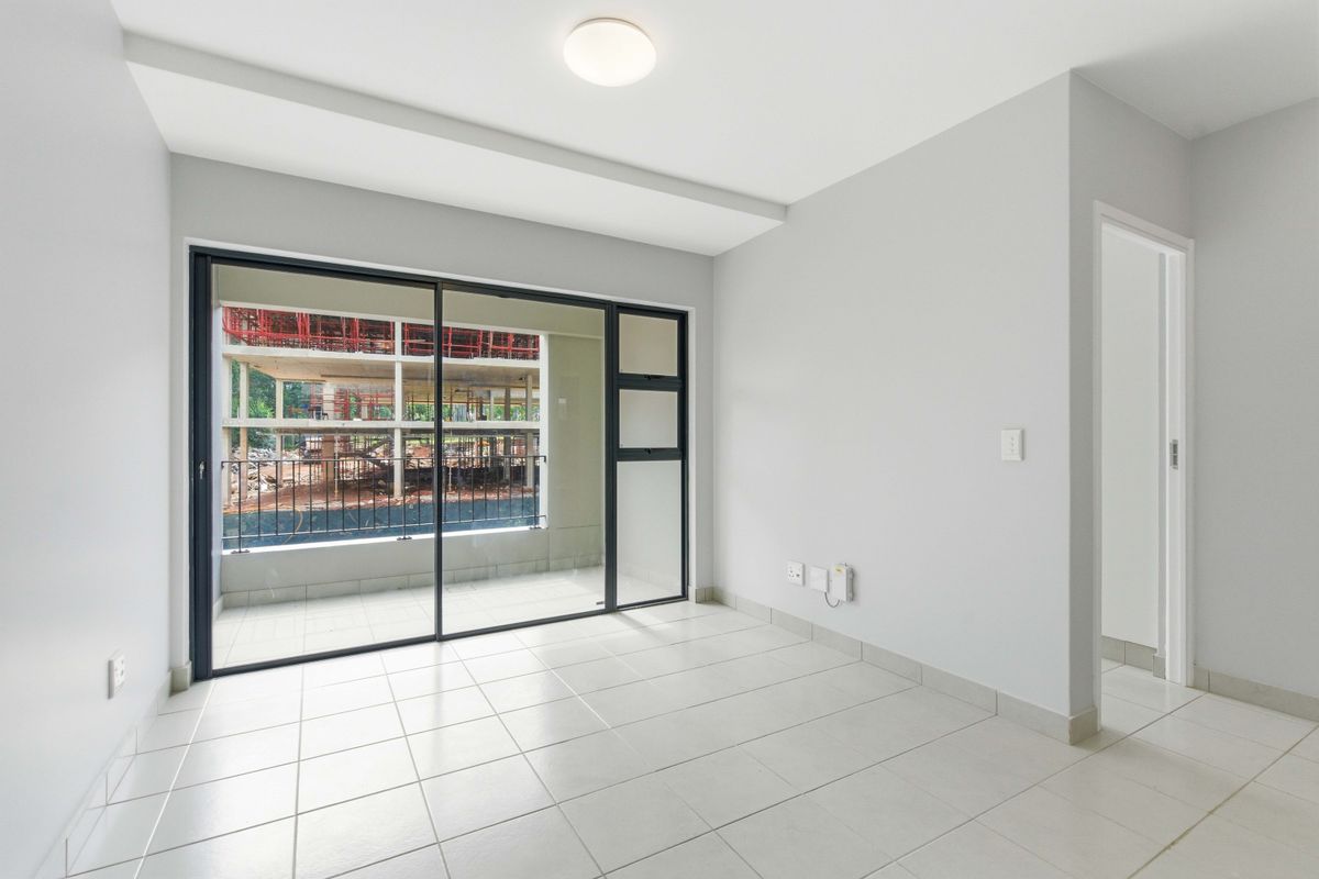 A bright, empty living area featuring neutral tiled flooring and a large sliding glass door that opens onto a balcony.