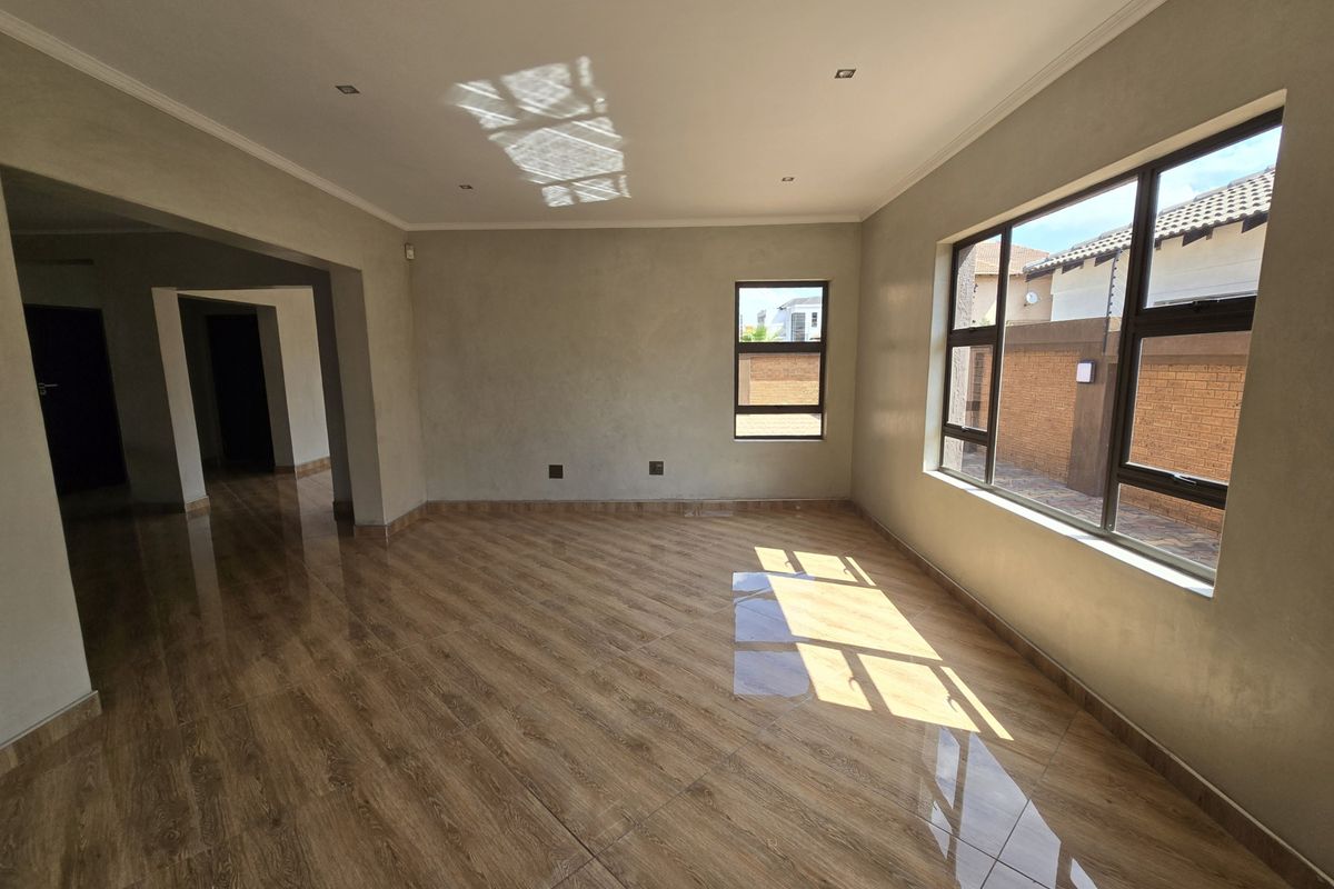 A bright, empty living room featuring modern wood-look tiled flooring, recessed downlights, and large aluminium-framed windows allowing for ample natural light.
