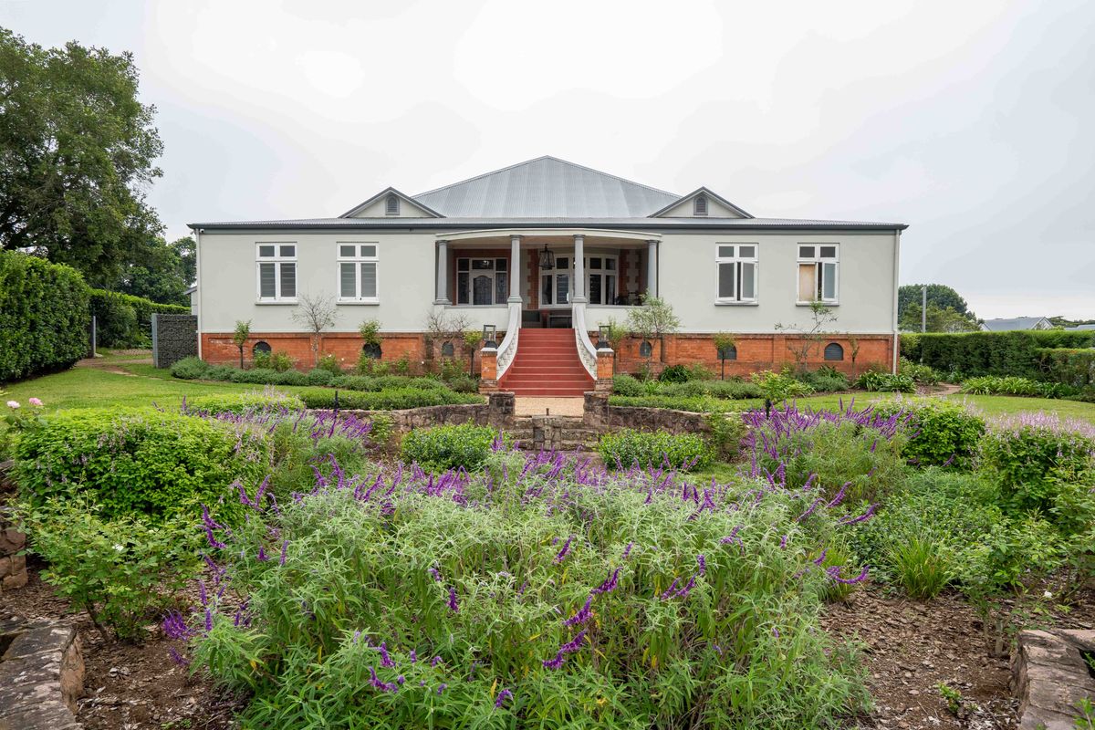 A well-maintained single-story residence featuring a covered veranda, brick foundation, and an expansive, beautifully landscaped garden with stone retaining walls.