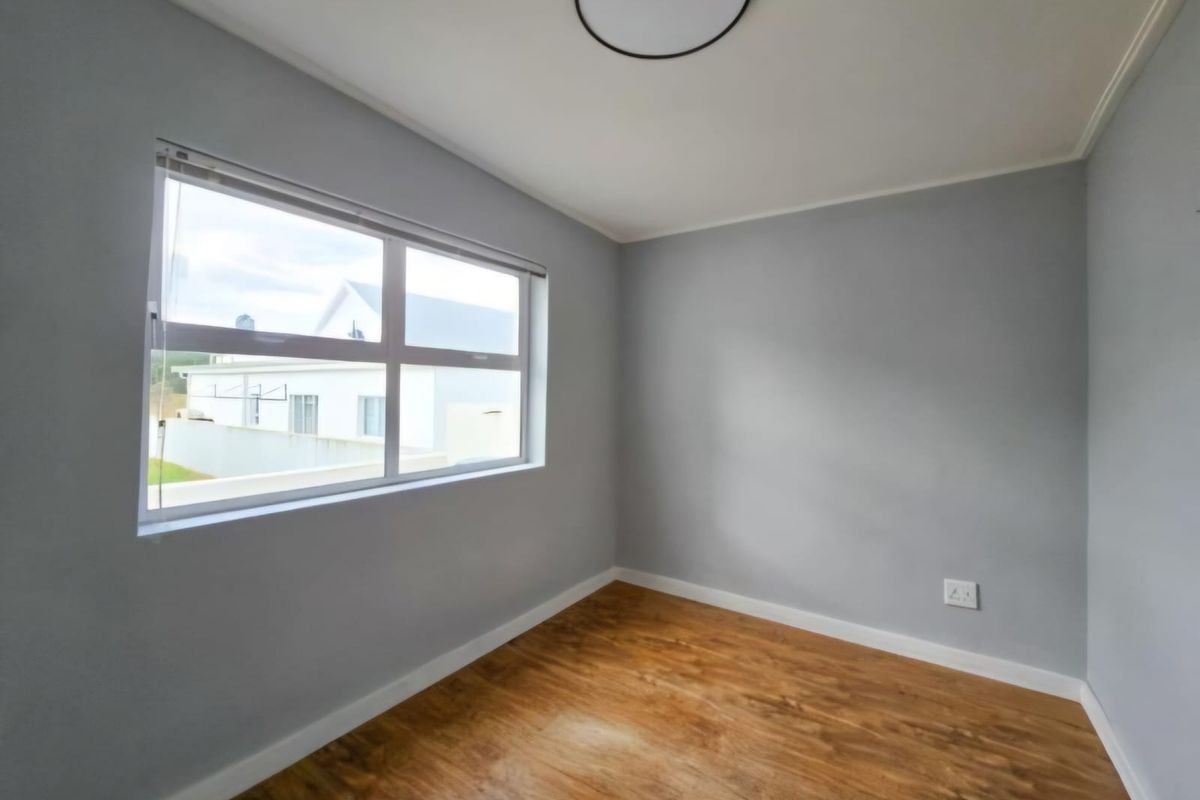 A clean, empty bedroom featuring light grey walls, warm-toned laminate flooring, and a large window providing natural light.
