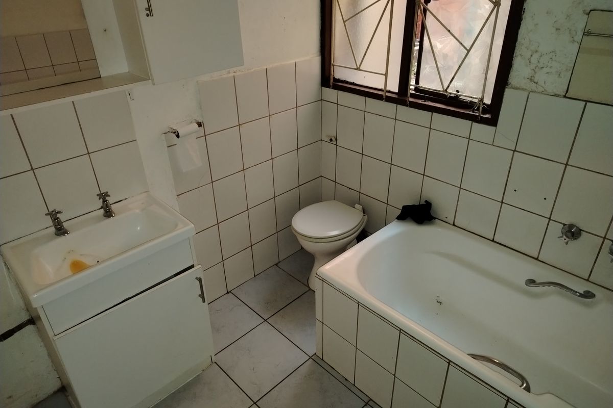 A dated bathroom featuring a standard bathtub, wall-mounted basin with vanity cabinet, and toilet, finished with white ceramic wall and floor tiles.