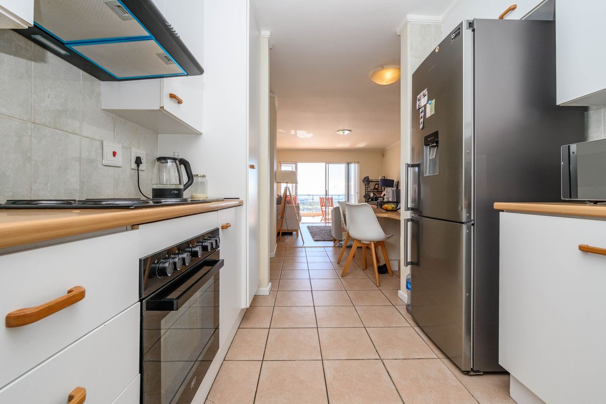 A functional galley-style kitchen featuring white cabinetry, laminate countertops, built-in oven, and hob, leading directly into an open-plan living area.