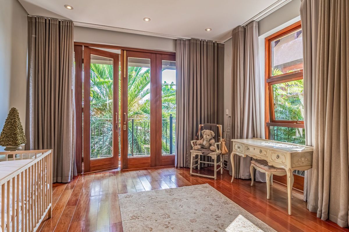 A bright bedroom featuring polished hardwood flooring, recessed ceiling lighting, and double wooden doors opening onto a private balcony with lush garden views.