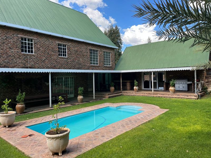 Spacious outdoor entertainment area featuring a swimming pool, brick-paved surround, and a covered patio with a built-in braai against a charming brick farmhouse exterior.